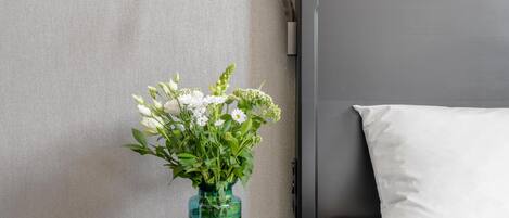 A bed with white bedding, a small table with a vase of flowers, and a wall-mounted lamp.