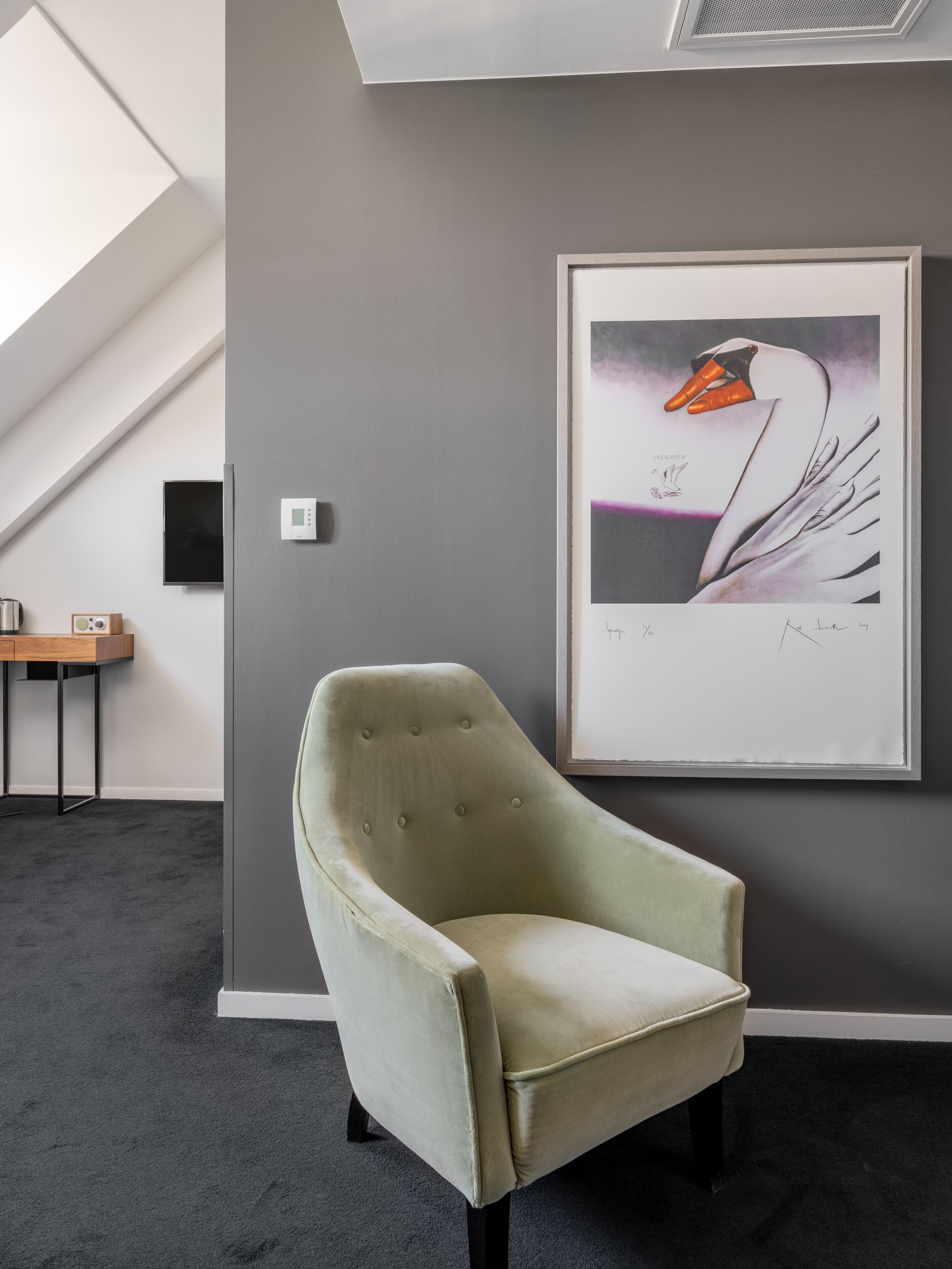 Una habitación moderna con pared gris, una obra de arte enmarcada de un cisne, un sillón verde, una mesita con un altavoz y un termostato.