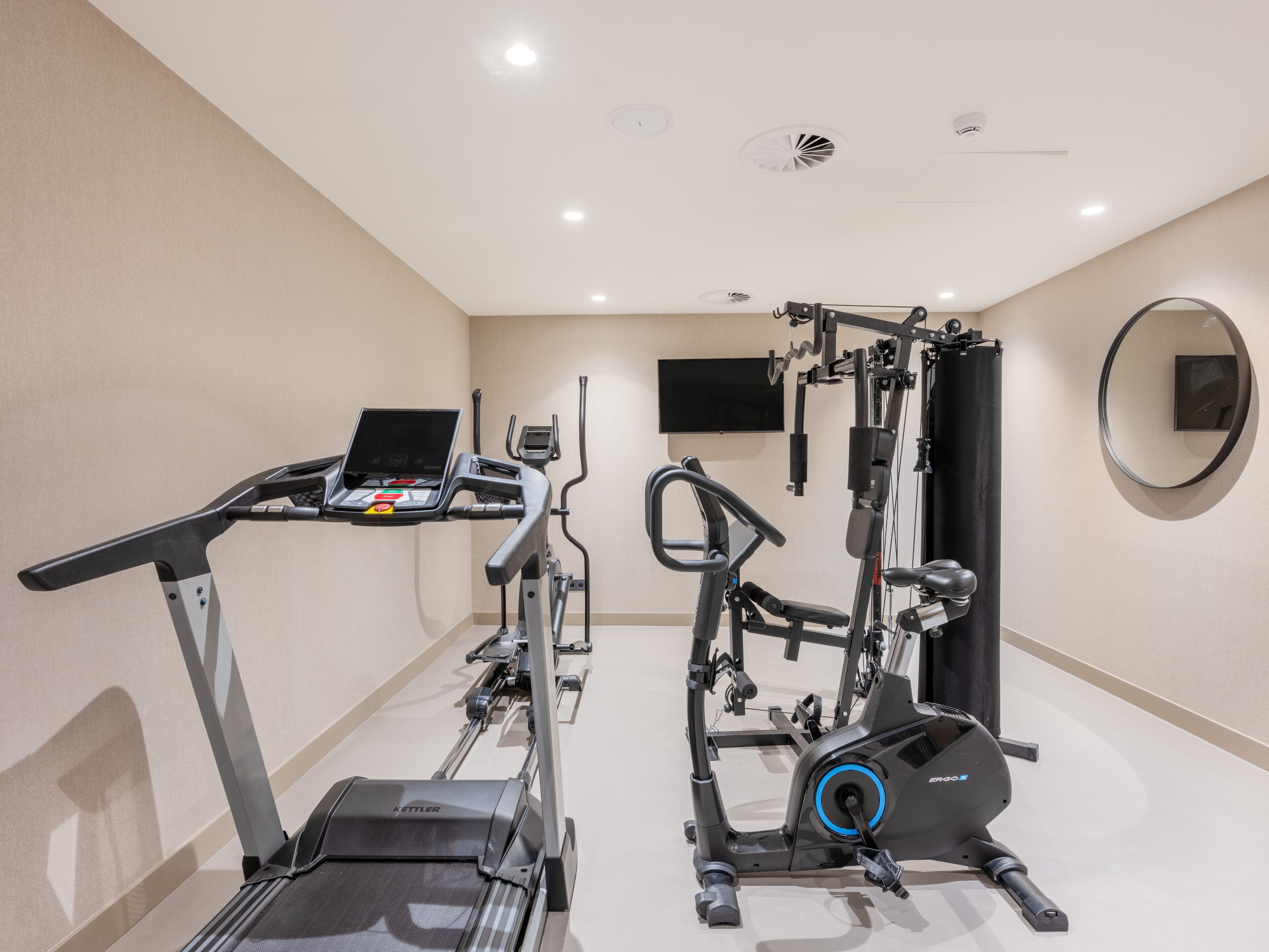 Een fitnessruimte met loopband, elliptische trainer en krachtapparaat, een televisie en een spiegel.