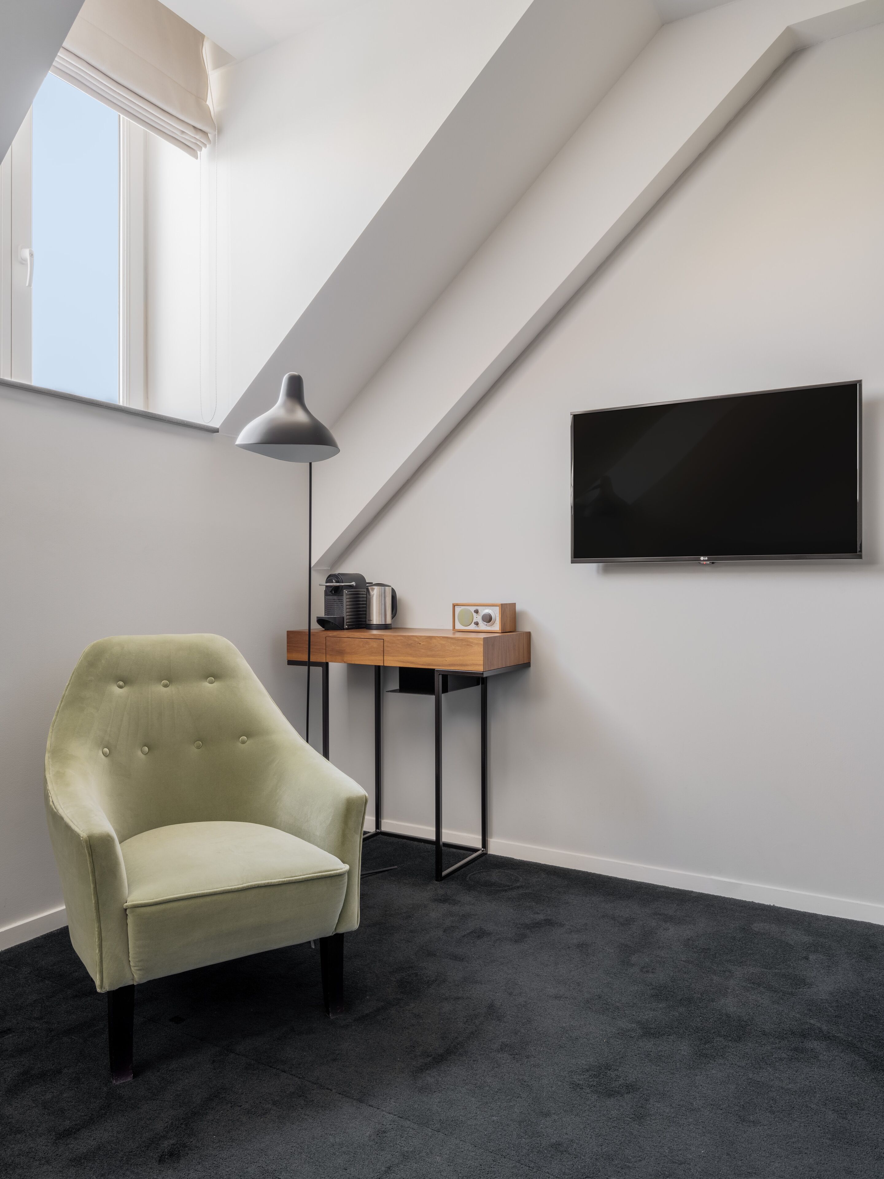 A room with a high ceiling, a large window, a flat-screen TV, a desk with a lamp, a chair, and a carpeted floor.