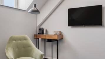 A room with a high ceiling, a large window, a flat-screen TV, a desk with a lamp, a chair, and a carpeted floor.