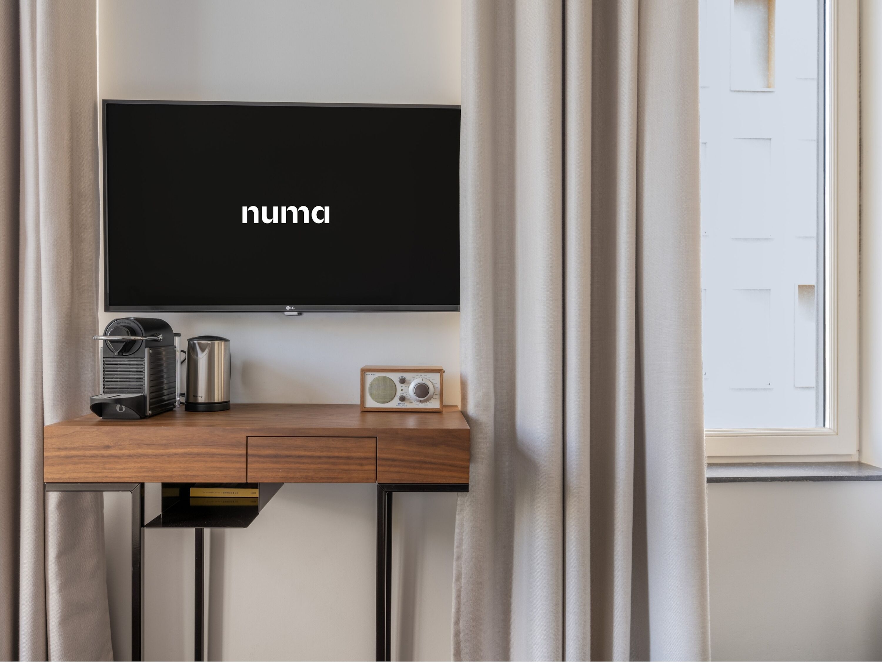 Una habitación moderna con televisión de pantalla plana montada en la pared, un escritorio de madera con cafetera, radio y una puerta con ventanal.