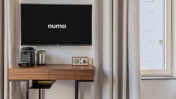Una habitación moderna con televisión de pantalla plana montada en la pared, un escritorio de madera con cafetera, radio y una puerta con ventanal.
