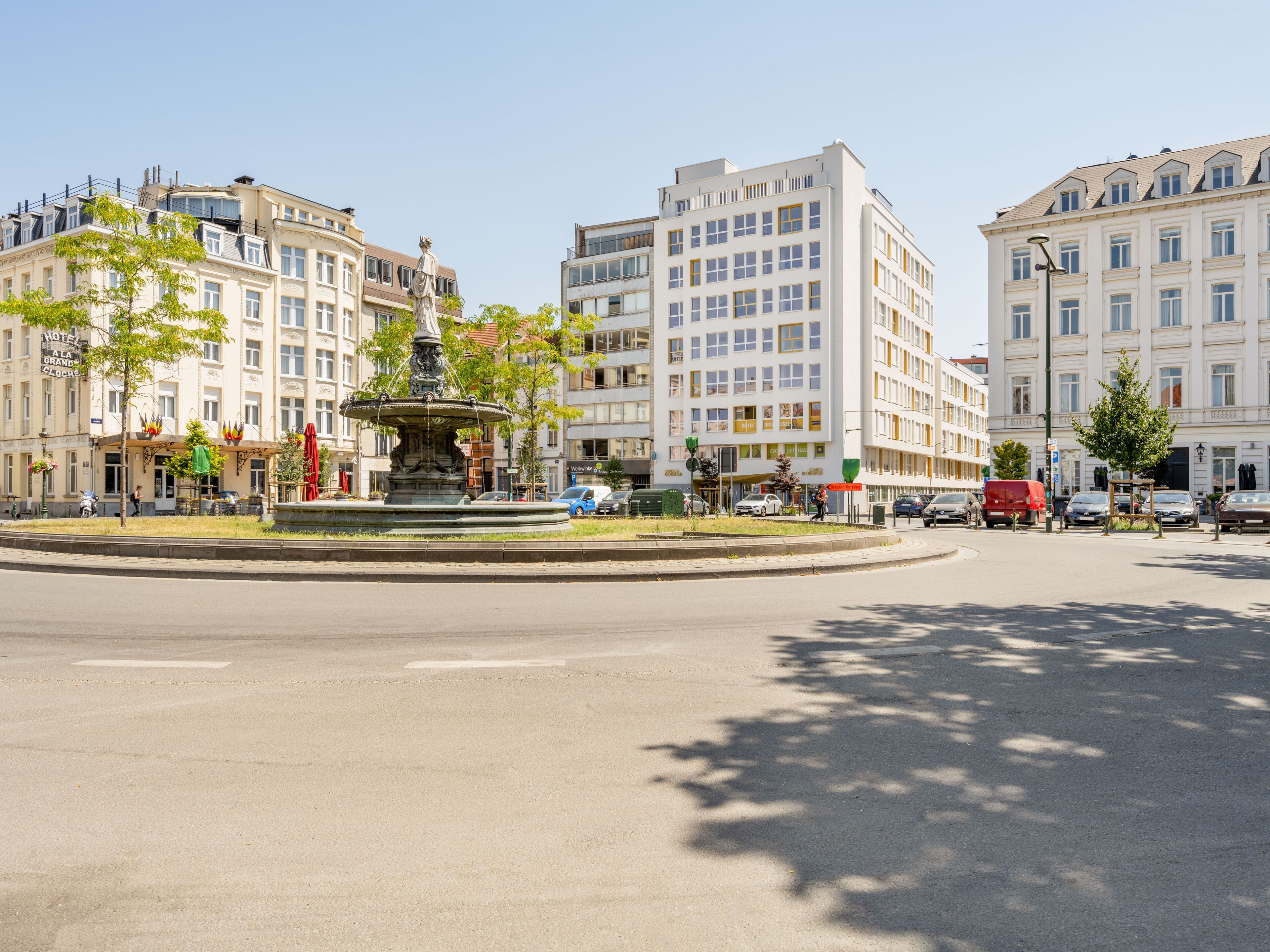 A public square with a central fountain, surrounded by multi-story buildings with commercial establishments on the ground floor.