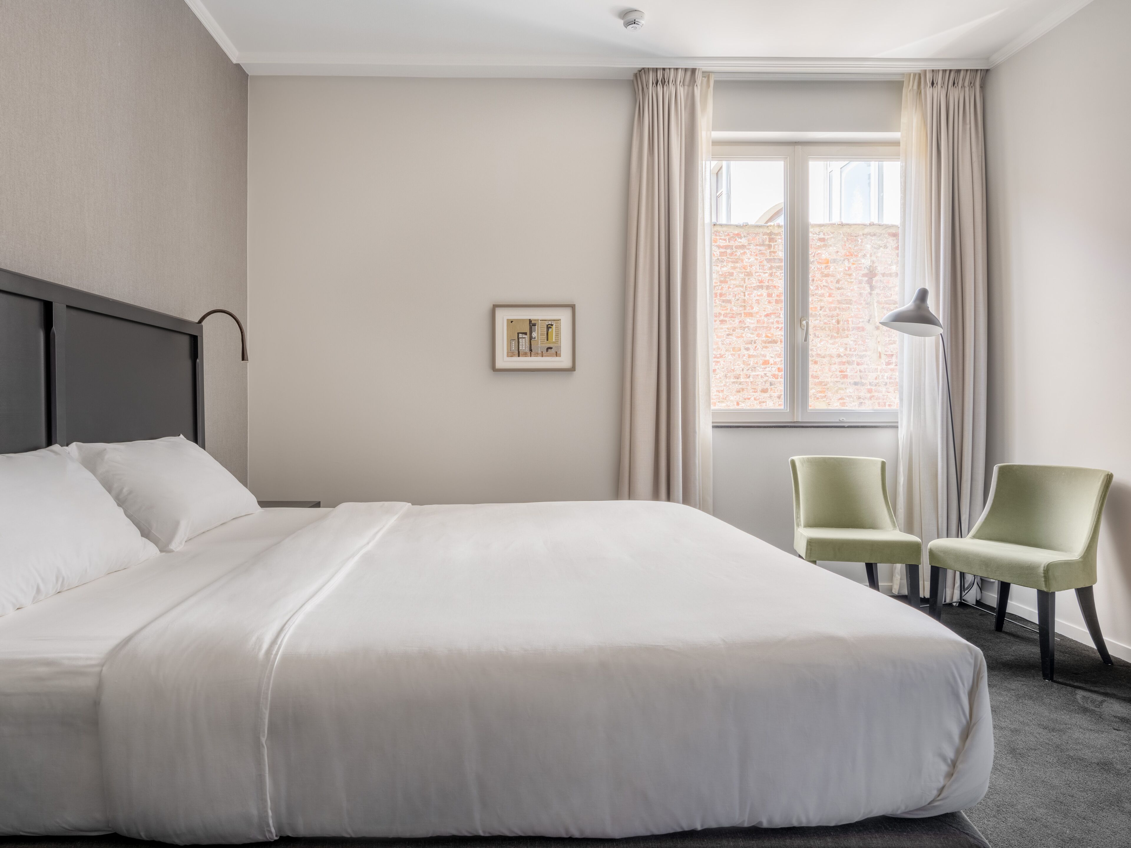 Une chambre d’hôtel avec un grand lit, deux chaises, une fenêtre avec des rideaux et un tableau encadré au mur.