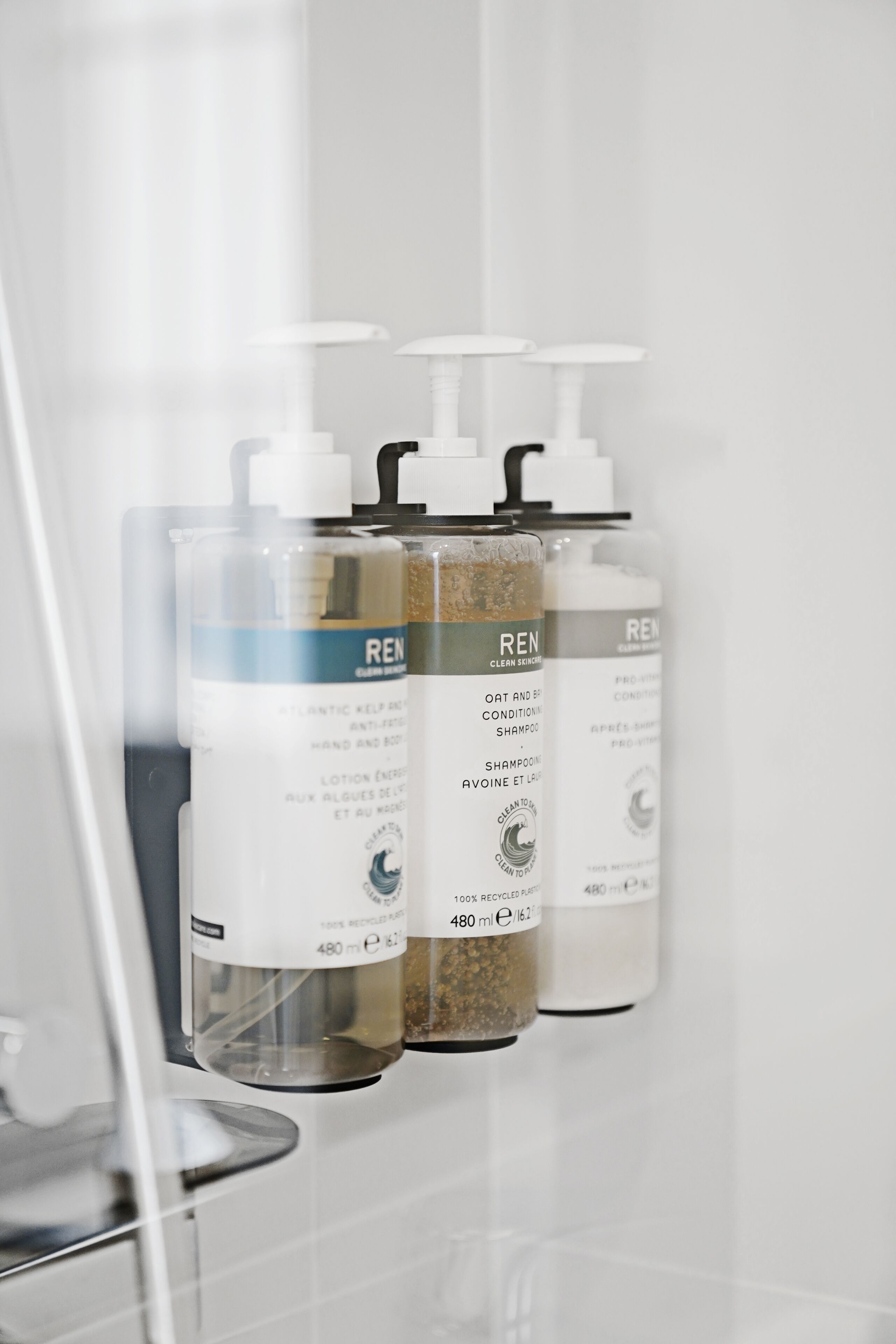 Trois bouteilles de shampoing et d’après-shampoing REN sont posées sur un mur de salle de bain.