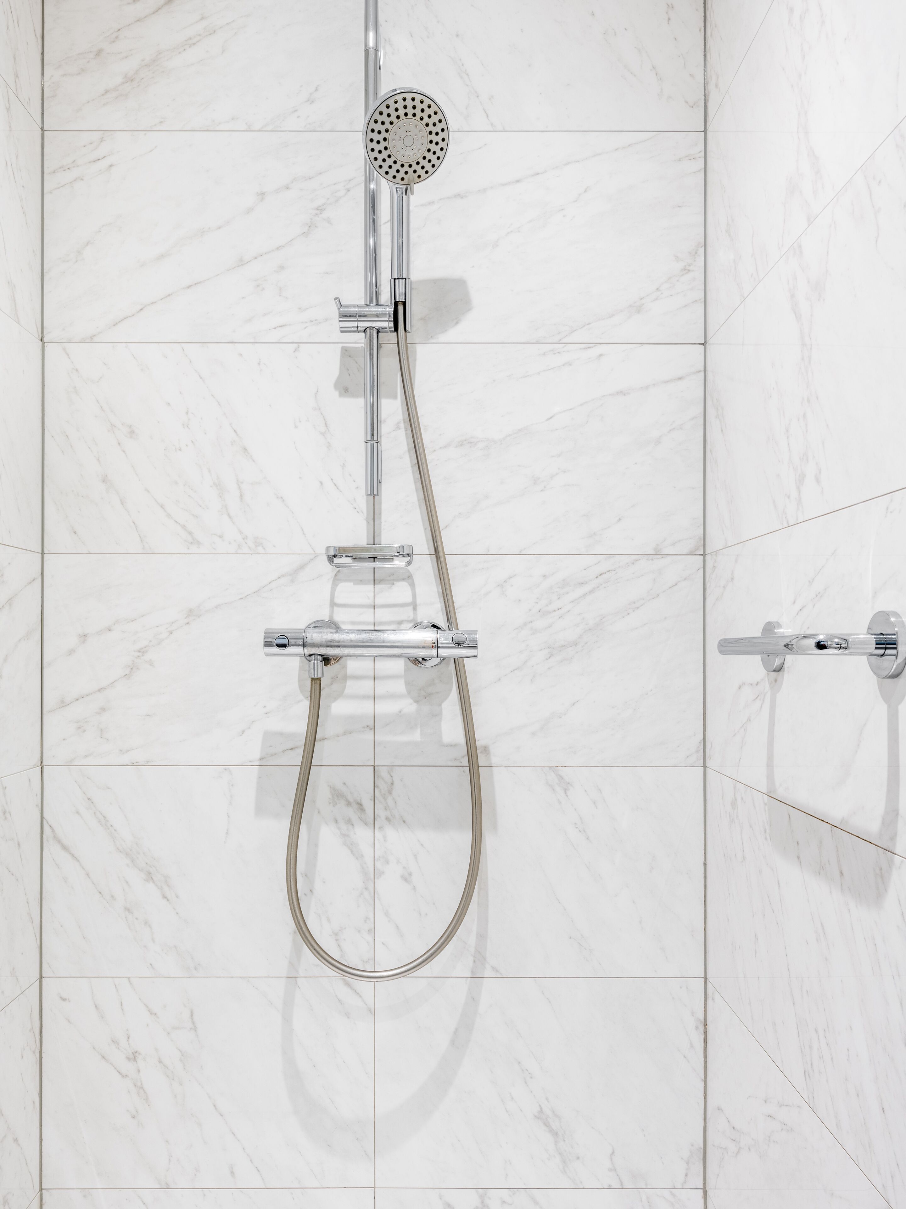 Ducha moderna con cabezal de ducha de mano y manguera flexible, montada en una pared con baldosas de mármol blanco.