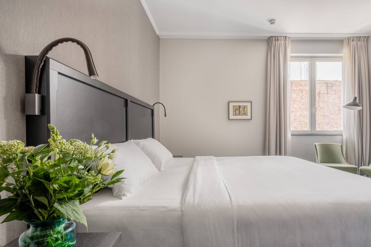 A hotel room with a large bed, a bedside table with a vase of flowers, a lamp, and a view of a brick building through the window.