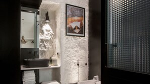 Un baño moderno con una pared blanca texturizada, un espejo grande, un lavamanos, un inodoro y una ventana con persianas.