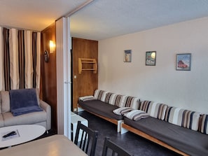 Une pièce compacte comprenant un canapé-lit, une petite table et une chaise. On y trouve deux lampes fixées au mur et une porte donnant sur une autre pièce.