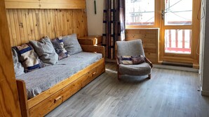 A cozy room with a wooden wall, a sofa with cushions, a chair, a small table, and a window with curtains. - Functional Studio for 4 · Ski-in/Ski-out at Plagne Villages (Plagne Villages)