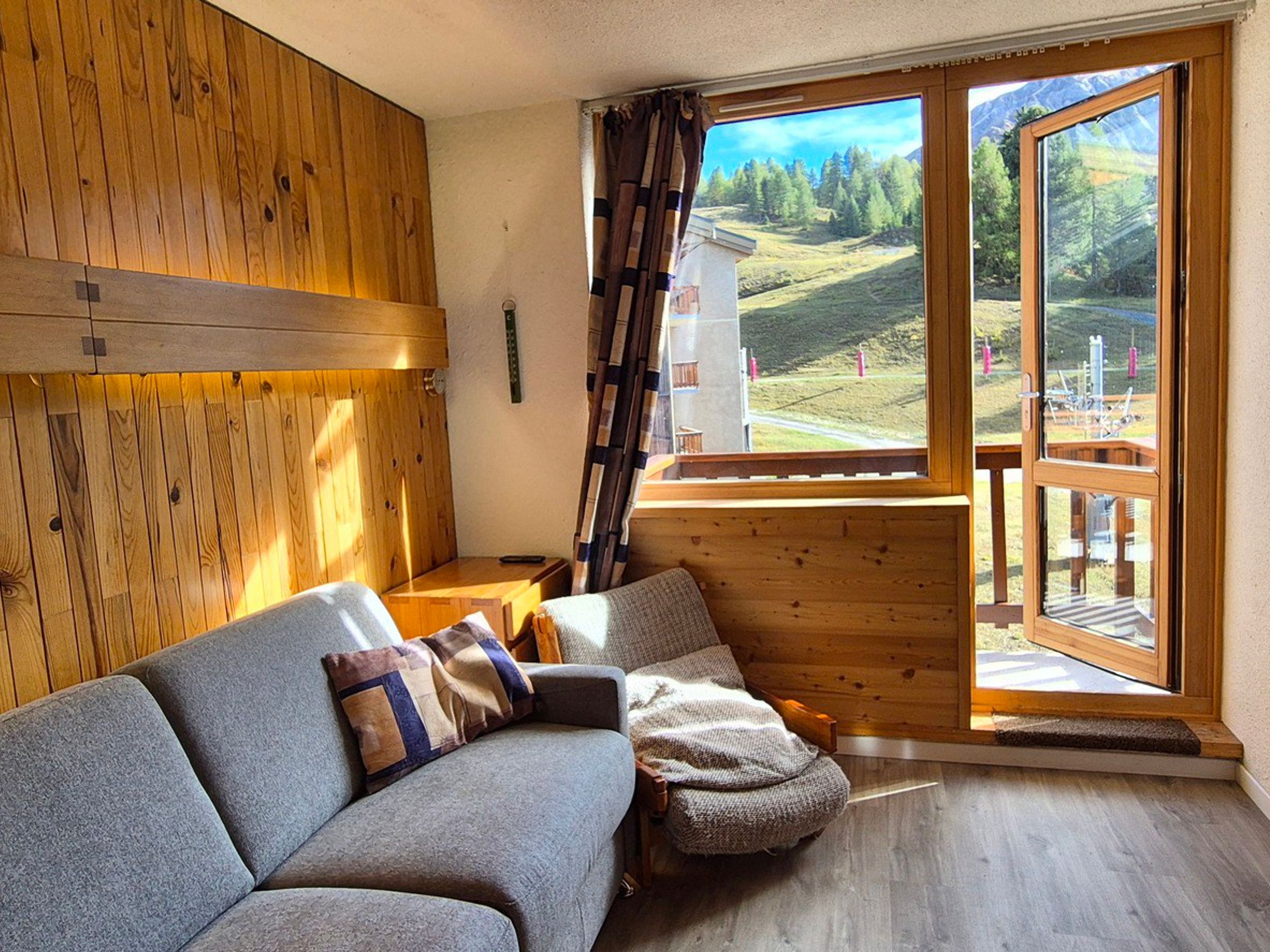 A cozy room with a wooden interior, a sofa with cushions, a window with a view of a mountainous landscape, and a wooden ladder leaning against the wall.