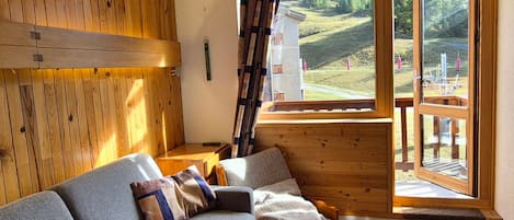 A cozy room with a wooden interior, a sofa with cushions, a window with a view of a mountainous landscape, and a wooden ladder leaning against the wall.