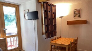 A cozy room with a wooden dining table and chairs, a television mounted on the wall, and a window with curtains. - Functional Studio for 4 · Ski-in/Ski-out at Plagne Villages (Plagne Villages)