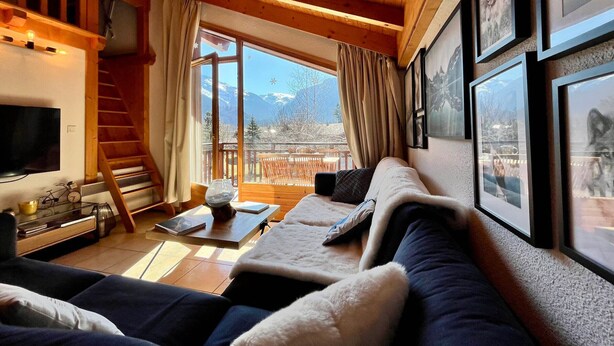 A cozy living room with a wooden ceiling, a large window with a mountain view, a television, and framed pictures on the wall.