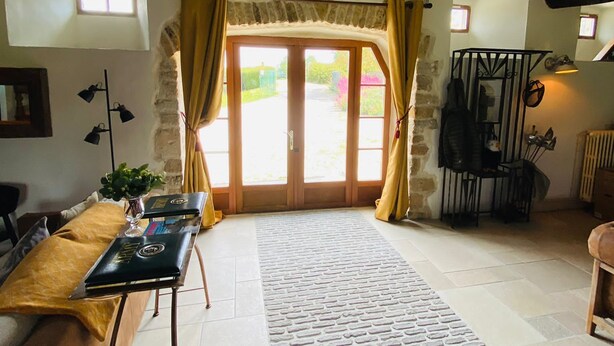 Un intérieur rustique avec des poutres apparentes, un mur en pierre, une grande table en bois recouverte d’une nappe jaune et un tapis à motifs menant à une porte ornée de rideaux.