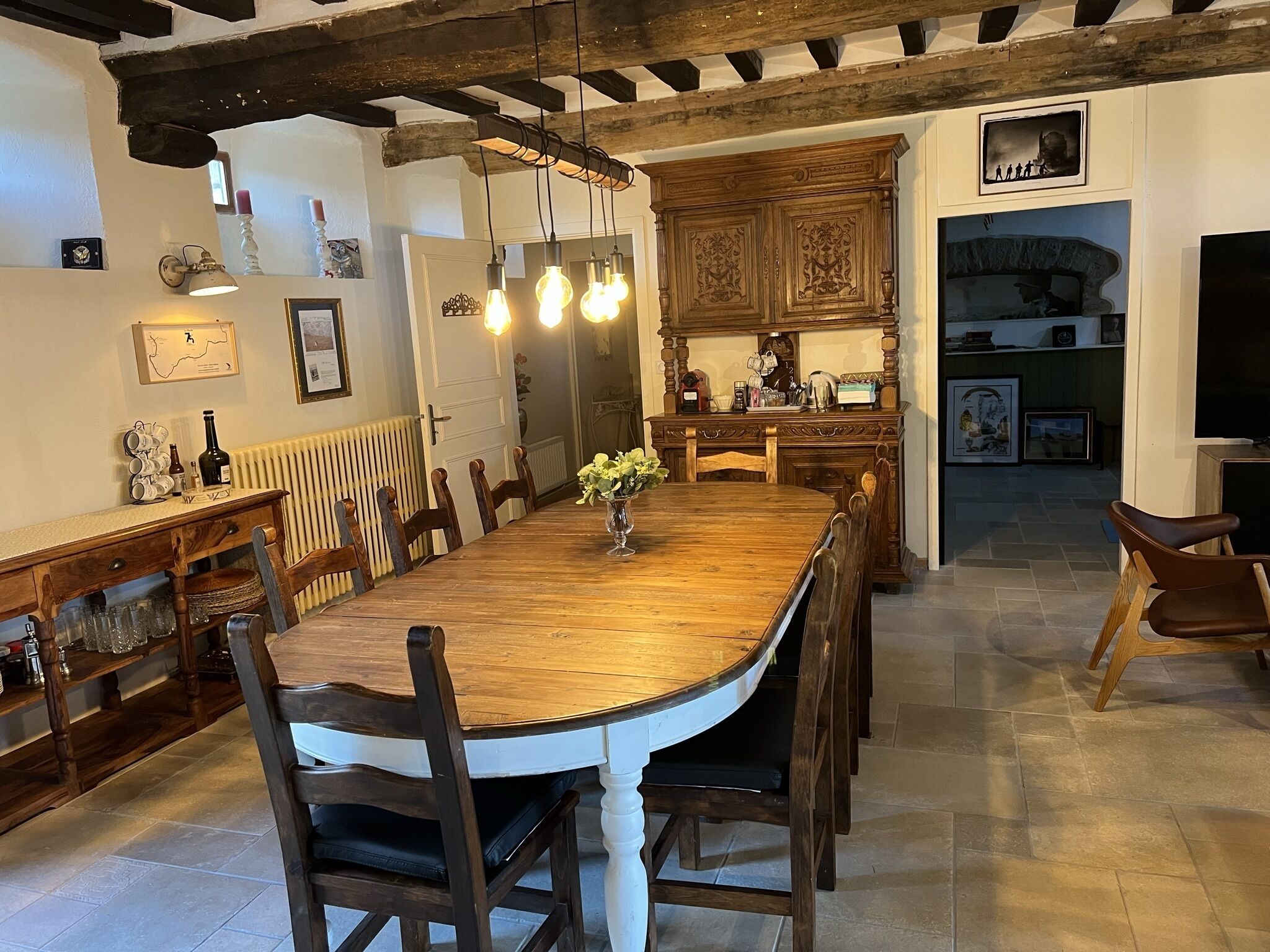 A dining area with a wooden table and chairs, a vase with flowers, and a kitchen in the background.