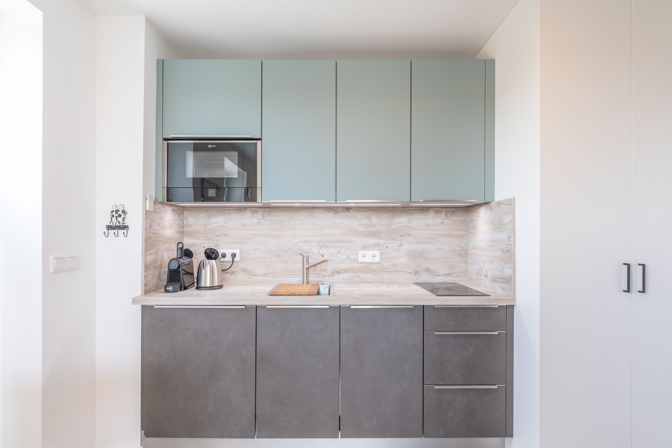 A compact kitchen with grey cabinets, a light blue upper cabinet, a stainless steel range hood, and a marble backsplash.