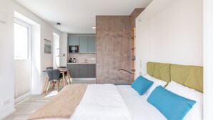 A modern hotel room with a large bed, a desk, and a kitchenette in the background.
