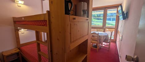 A bunk bed with a wooden headboard, a small table with chairs, and a window with a view of a green landscape.