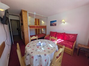 A room with a bunk bed, a round table with chairs, a red sofa, and a wall-mounted TV.