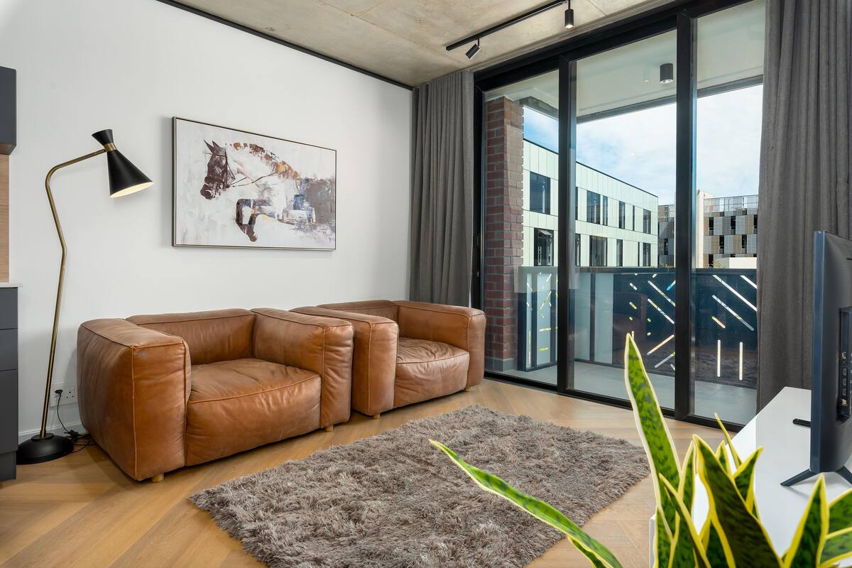 A modern living room with a brown leather sofa, a gray shag rug, a floor lamp, and a large window with a city view.