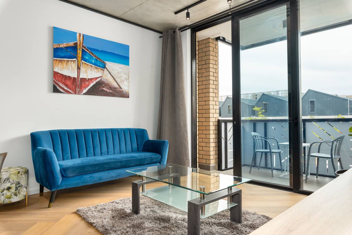 A modern living room with a blue sofa, a glass coffee table, and a view of a balcony with outdoor furniture.