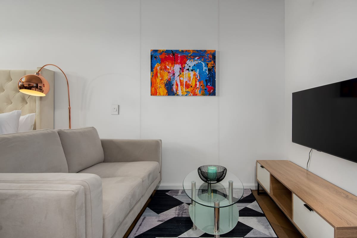 A modern living room with a beige sofa, a glass coffee table, a colorful abstract painting, a black TV, and a wooden TV stand.