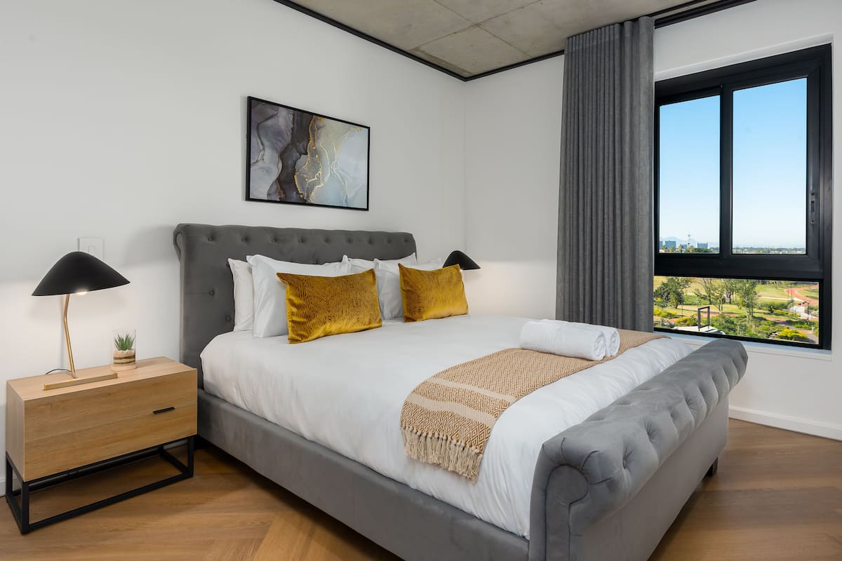 A modern bedroom with a large bed, a bedside table, a lamp, a painting, and a window with a city view.