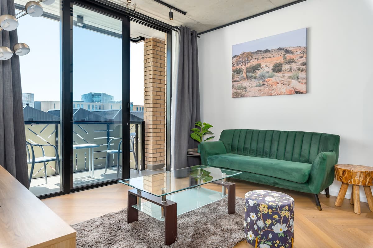 A modern living room with a glass coffee table, a green sofa, and a view of a cityscape through large sliding doors.