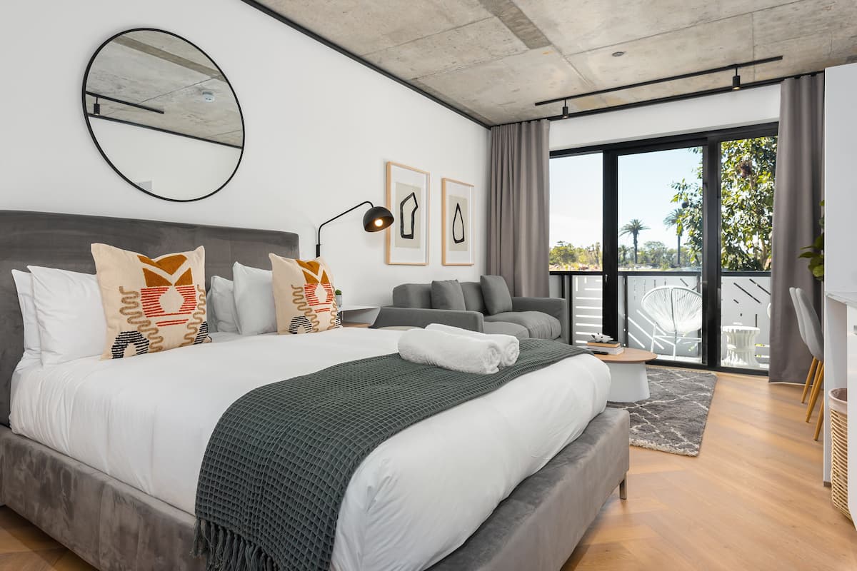 A modern hotel room with a large bed, a round mirror, and a balcony with a view of palm trees.