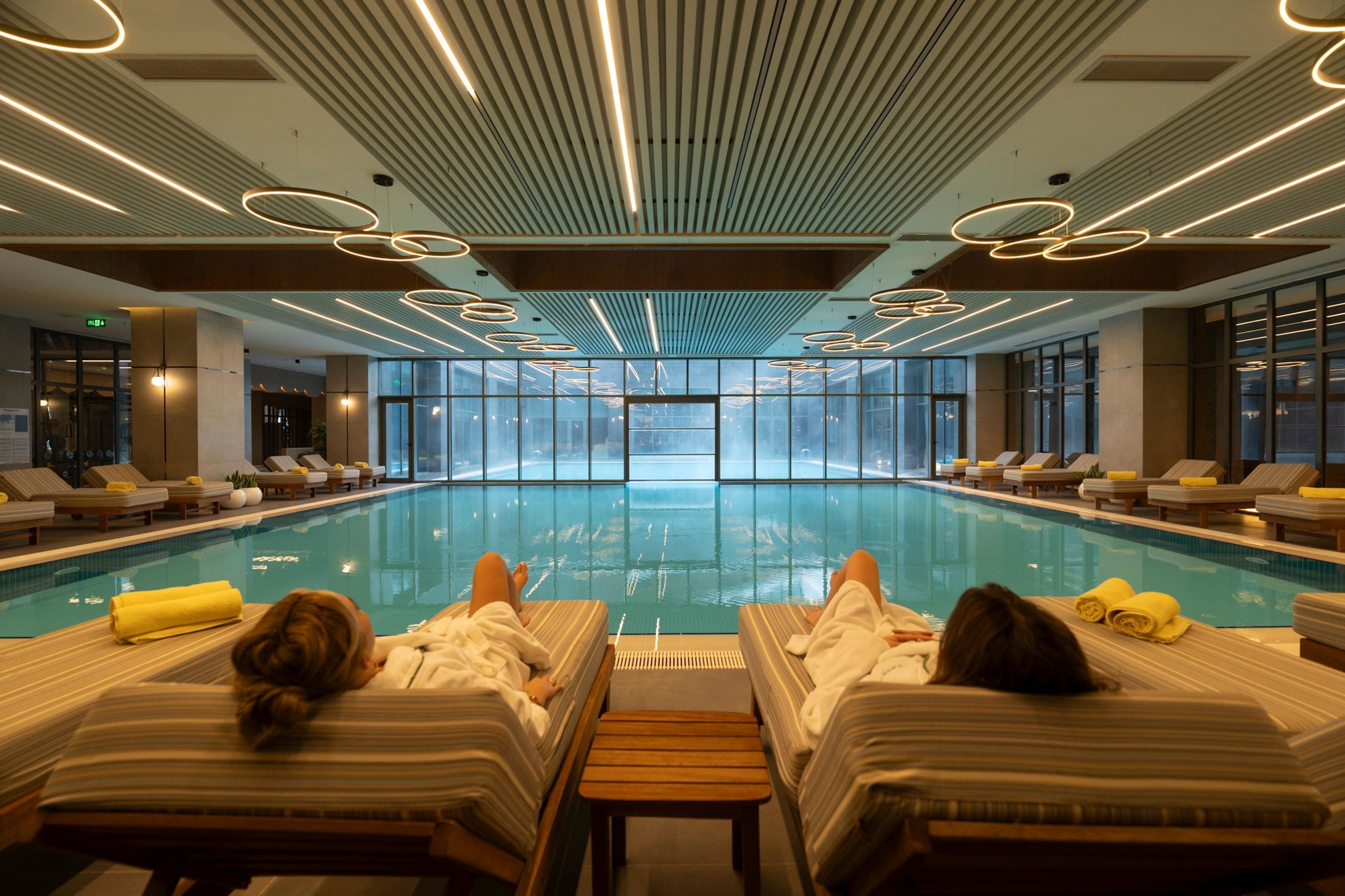 A large indoor swimming pool with lounge chairs and towels, surrounded by large windows.
