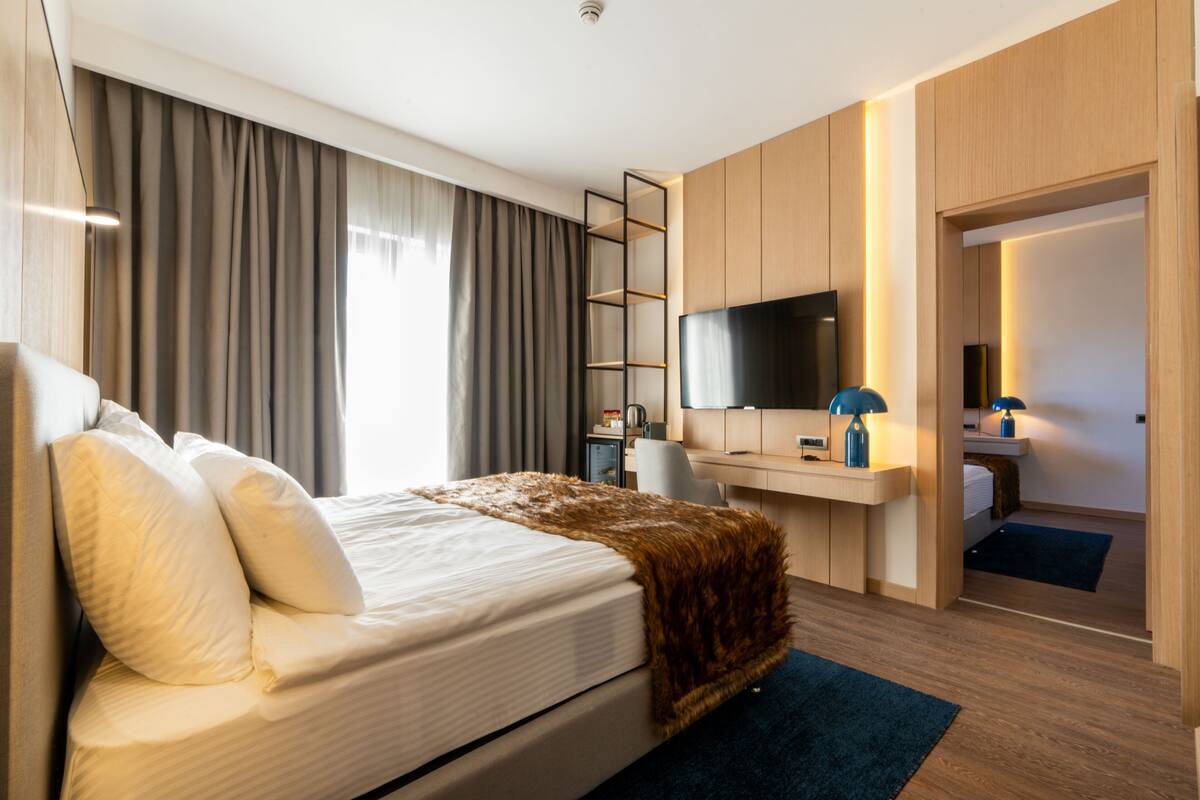 A modern hotel room with a large bed, a desk with a computer, a TV, and a wardrobe.