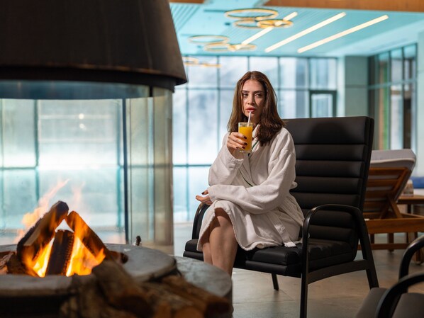A person in a white bathrobe sitting by a fireplace in a modern indoor setting with glass walls and a lounge area. - Megasaray Mount Erciyes (Kayseri)