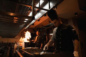 A chef preparing food in a kitchen with a visible fire, stainless steel appliances, and a high ceiling. - Megasaray Mount Erciyes (Kayseri)