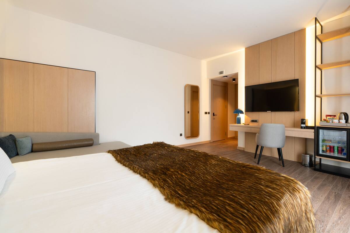 A modern hotel room with a large bed, a desk, a television, and a minibar.