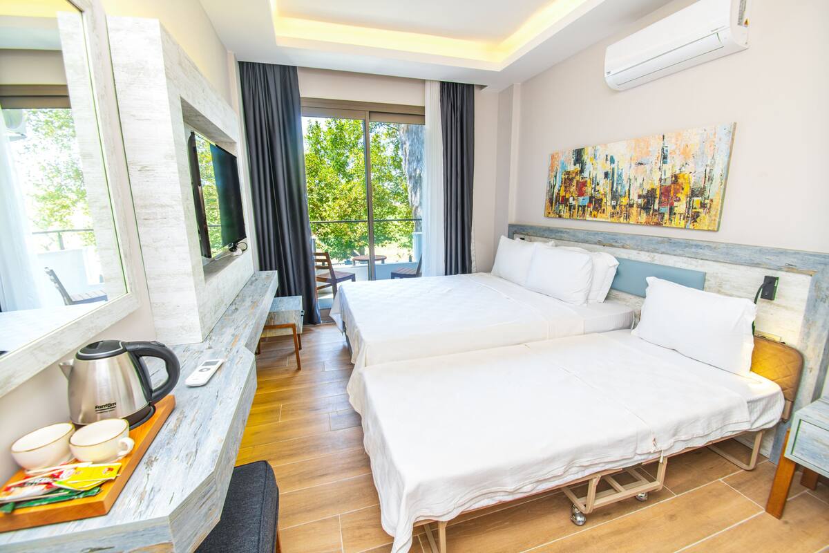 A hotel room with two beds, a TV, a painting, and a balcony with a view of trees.