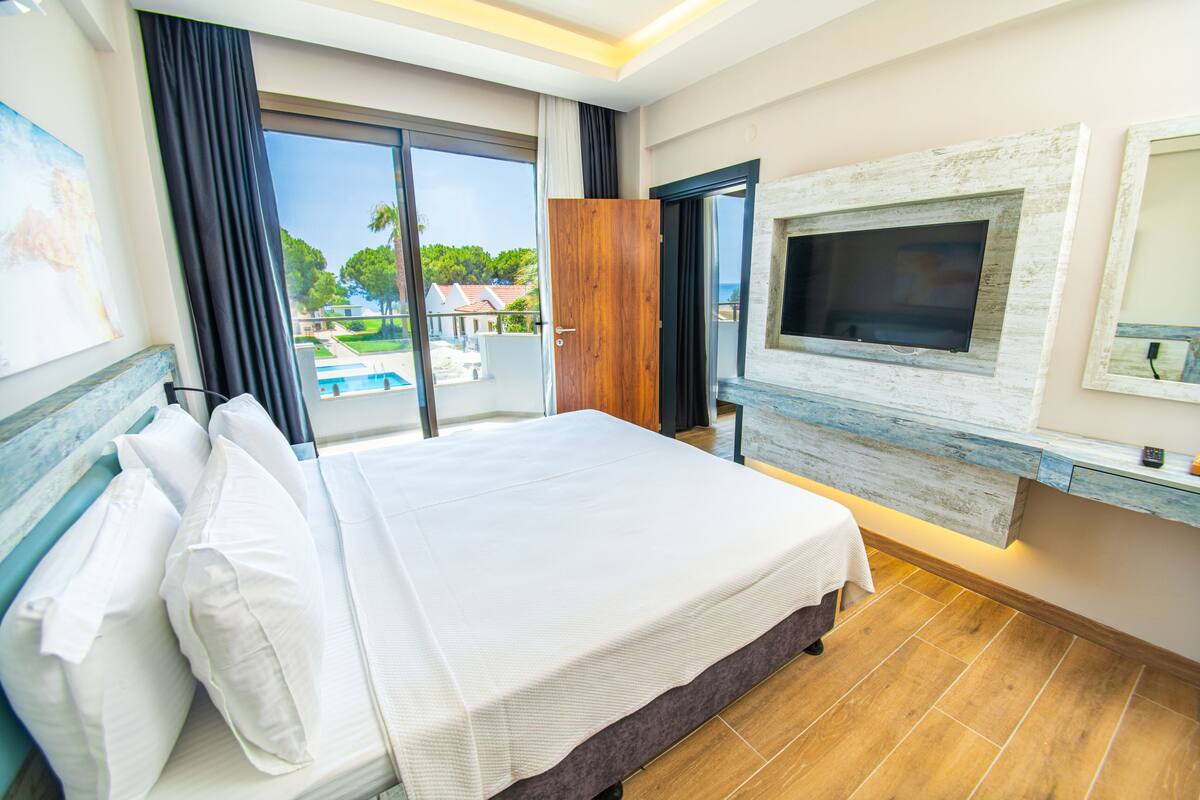 A hotel room with a large bed, a TV mounted on a rustic wall, a sliding glass door to a pool, and a view of the surrounding landscape.