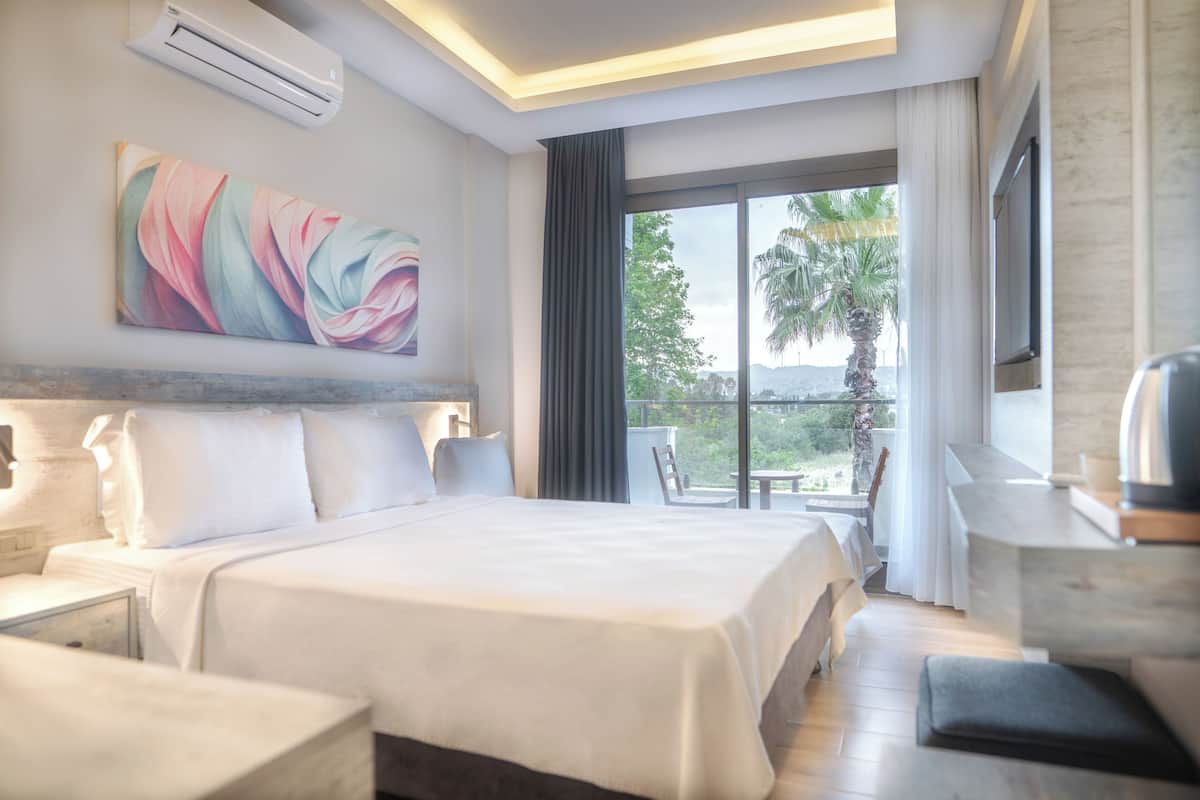 A hotel room with a large bed, a desk, a chair, a TV, and a balcony with a view of palm trees.