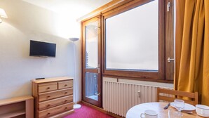 Et hotelværelse med en trækommode, et fjernsyn ophængt på væggen, et spisebord med et rødt tæppe og et stort vindue med gardiner.