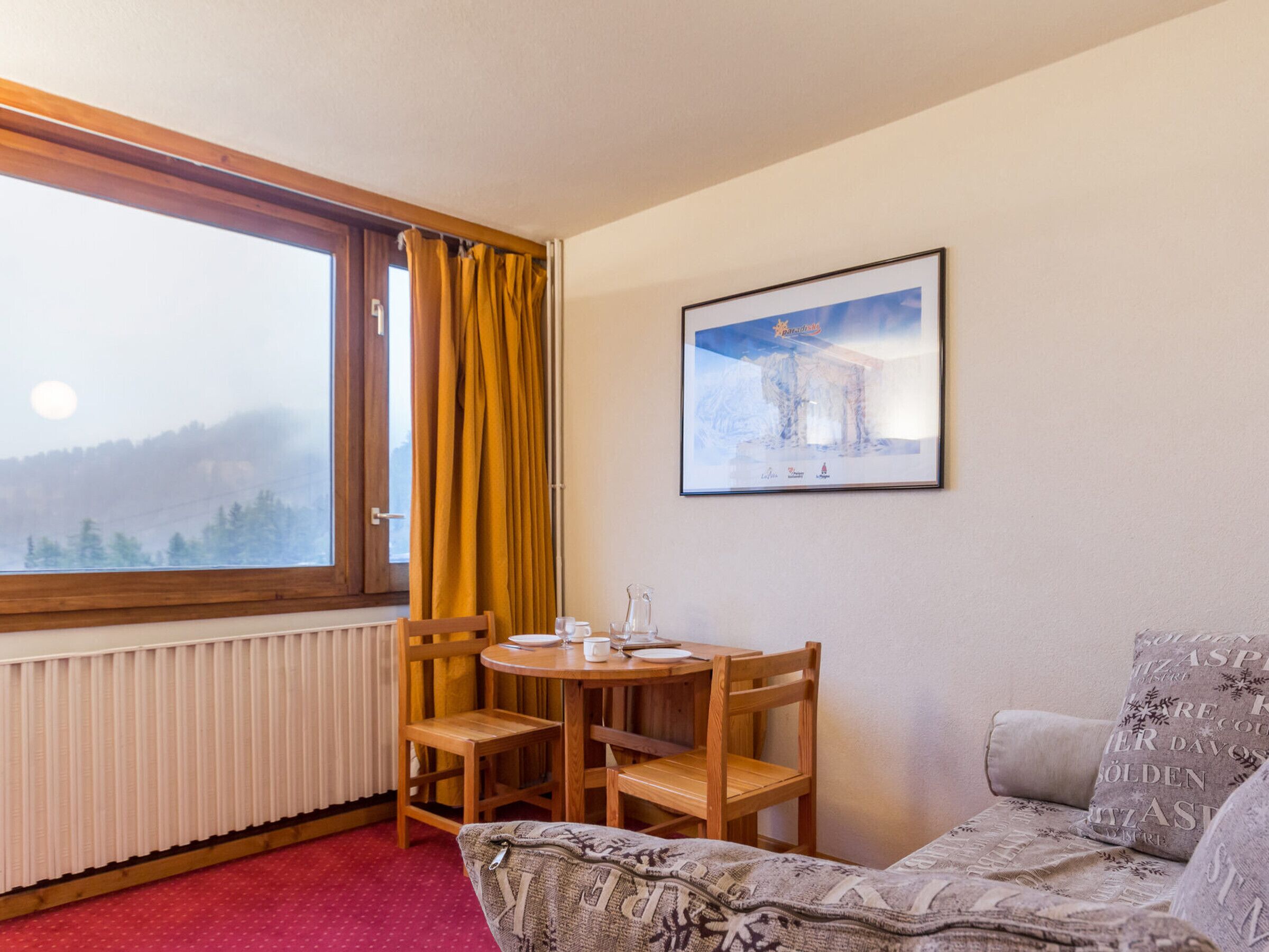 A cozy room with a wooden table, chairs, a sofa, a window with curtains, and a framed picture on the wall.