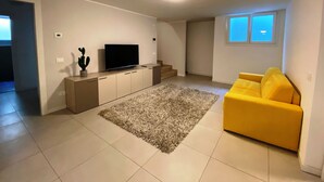 A living room with a flat-screen TV on a wooden cabinet, a yellow sofa, and a grey shag rug. - Villa Silvia, with private heated pool and garden. (Solarolo)