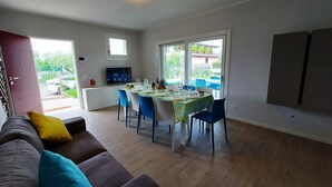 A modern living room with a dining area, wooden flooring, a sofa with colorful cushions, a TV, and a kitchen area with a window. - Villa Silvia, with private heated pool and garden. (Solarolo)