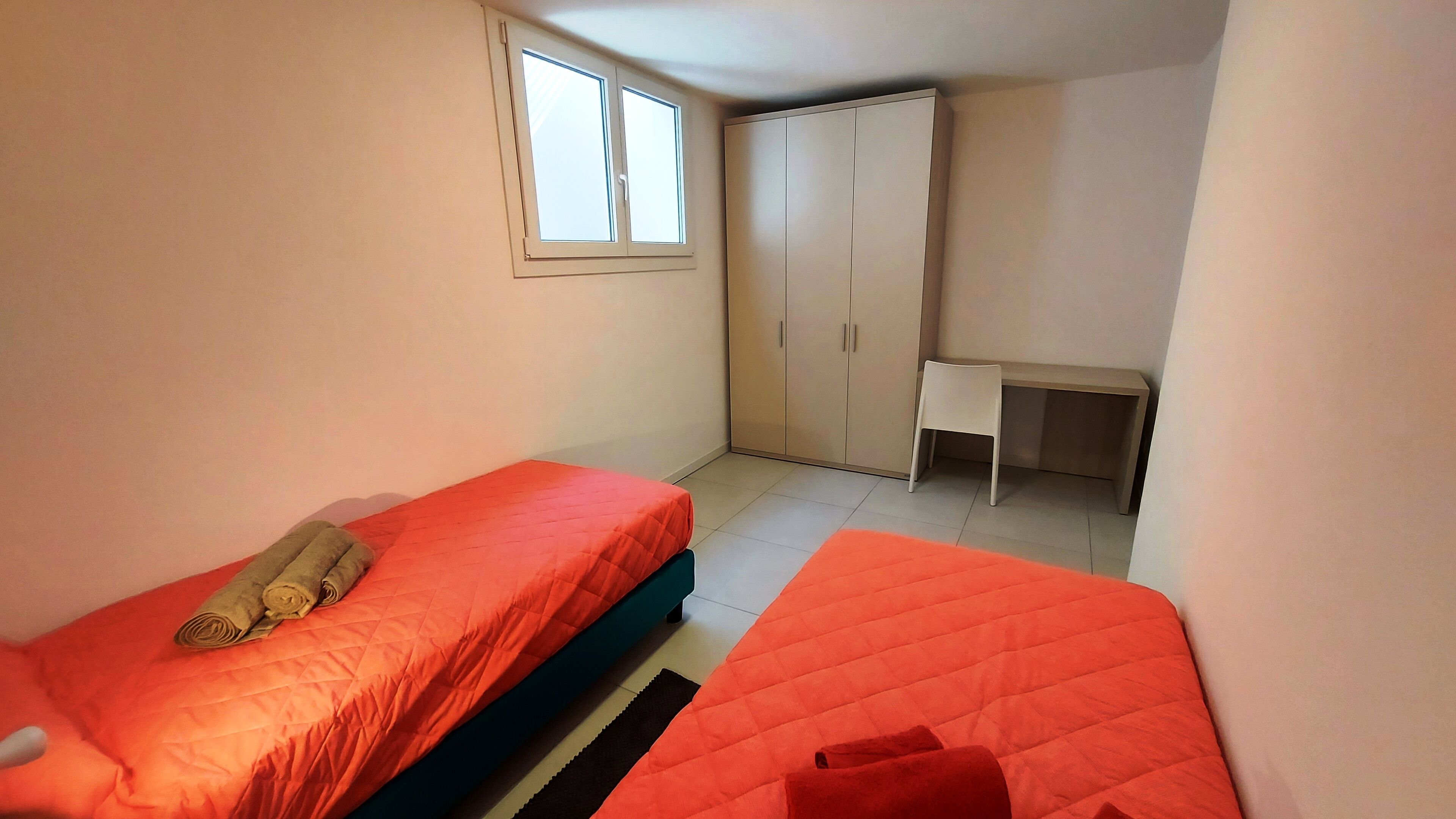 A small, well-lit room with two beds, a desk, and a wardrobe.
