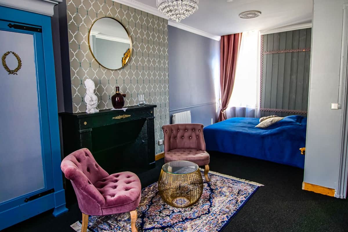 Quarto com cama, penteadeira com espelho, cômoda, poltrona rosa e lustre.
