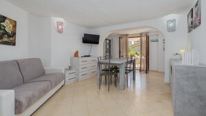 Un salon avec un canapé gris, une table à manger avec des chaises, un téléviseur fixé au mur et un coin cuisine visible à travers une arche.