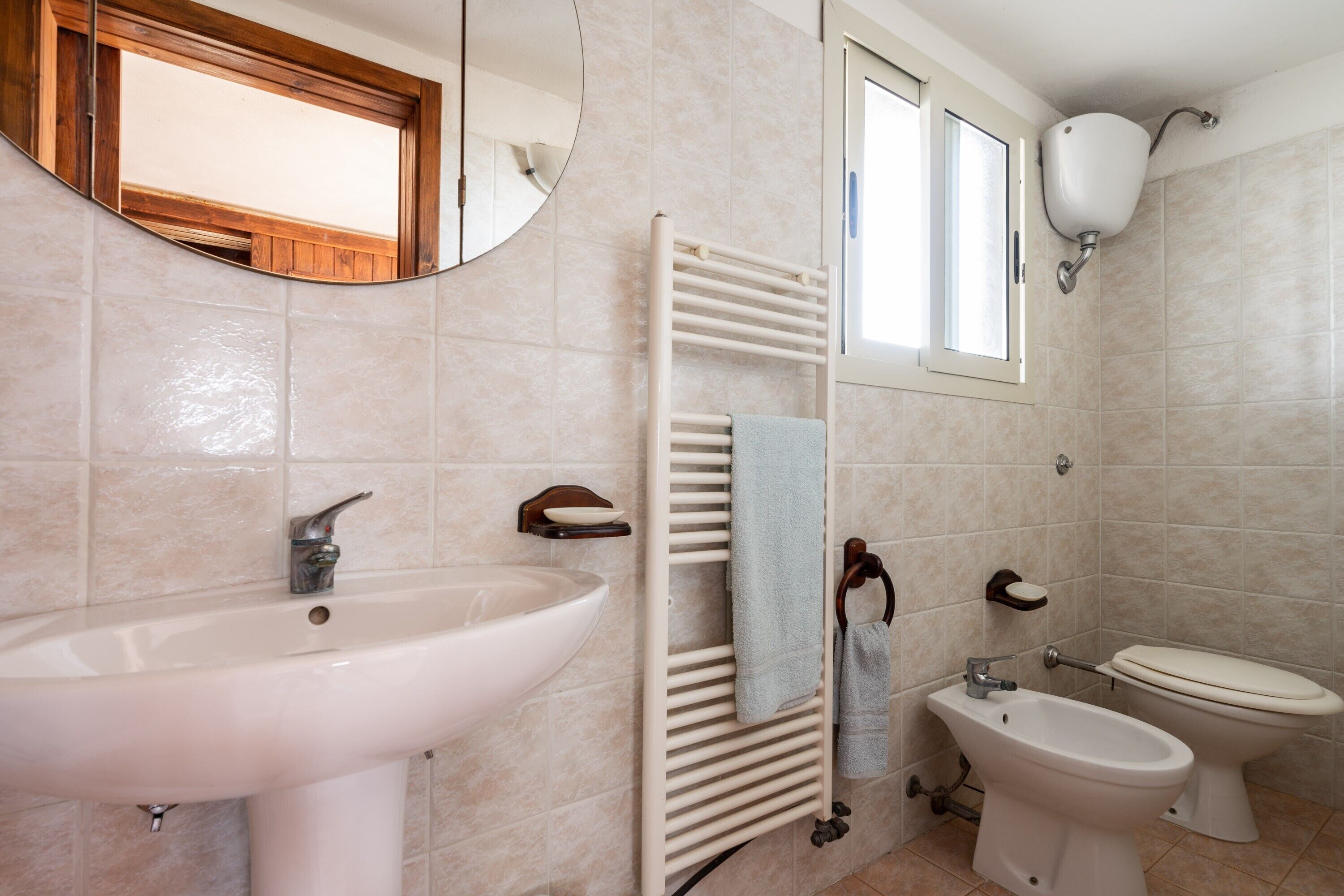 A bathroom with a sink, bidet, toilet, towel rack, mirror, and window.