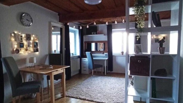 A spacious living room with a wooden ceiling, a dining area with a table and chairs, a bookshelf, and a large window providing natural light. - Cozy fresh apartment with rustic roof in quiet area (Graz)