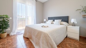 Un dormitori amb llit, mesetes de nit, una planta en test i una finestra amb cortines primes.