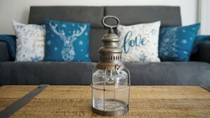 A clear glass lantern on a wooden table with a grey sofa and blue floral pillows in the background. - The Family Lake - Appartement 2 Chambre, Terrasse & Parking (Lathuile)